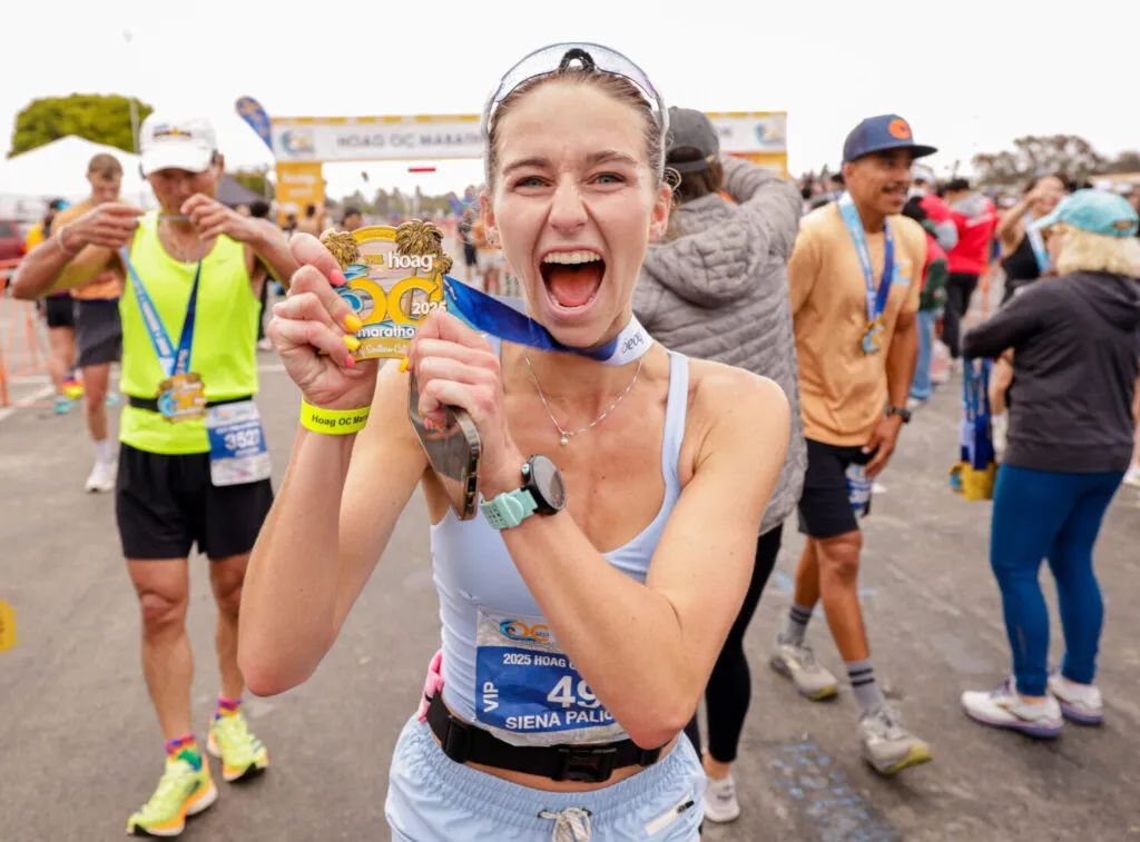 A jubilant female runner holds up her marathon medal, mouth open in a shout of excitement, with other runners and a finish line banner in the background.