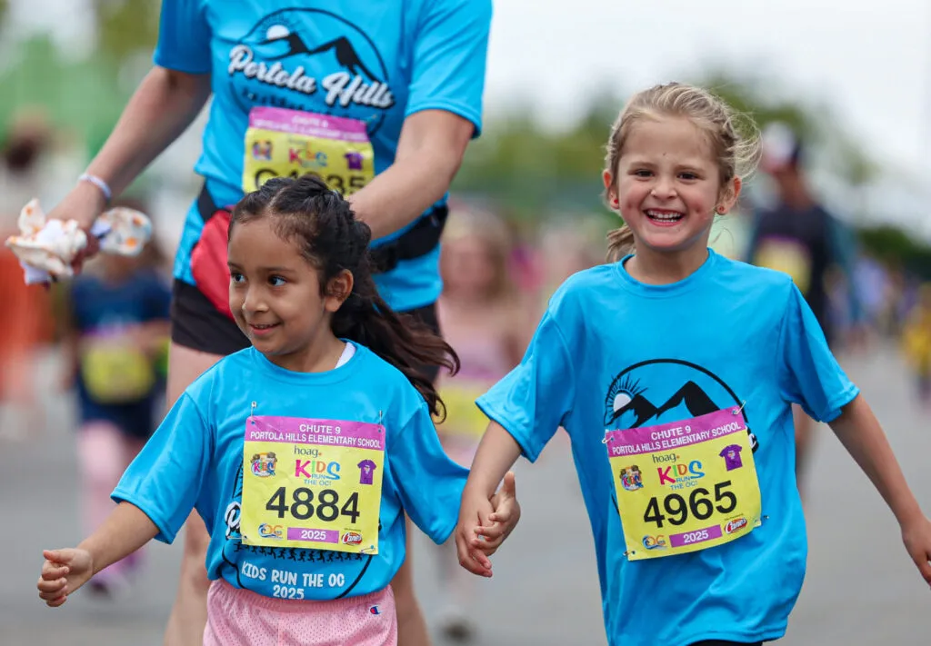 Two young girls in blue race t-shirts, smiling and holding hands as they run.