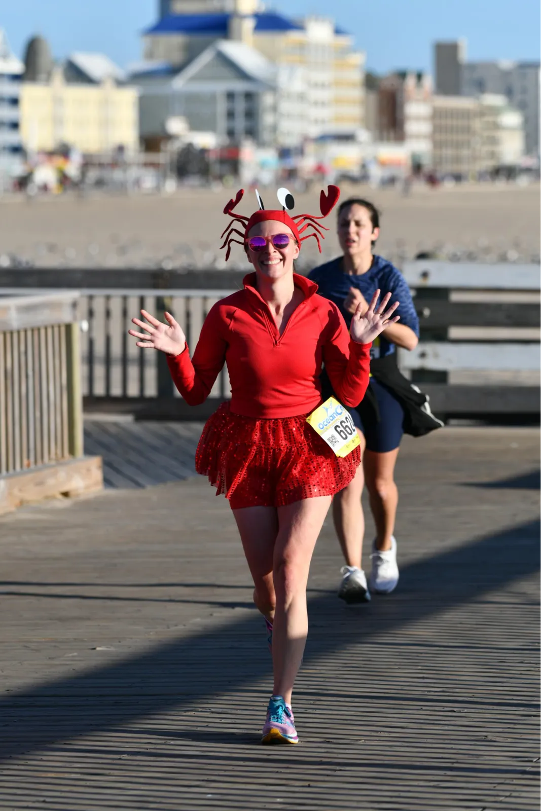 A runner wearing a crab costume and sunglasses smiles and waves while running on a boardwalk, with other runners and buildings in the background.