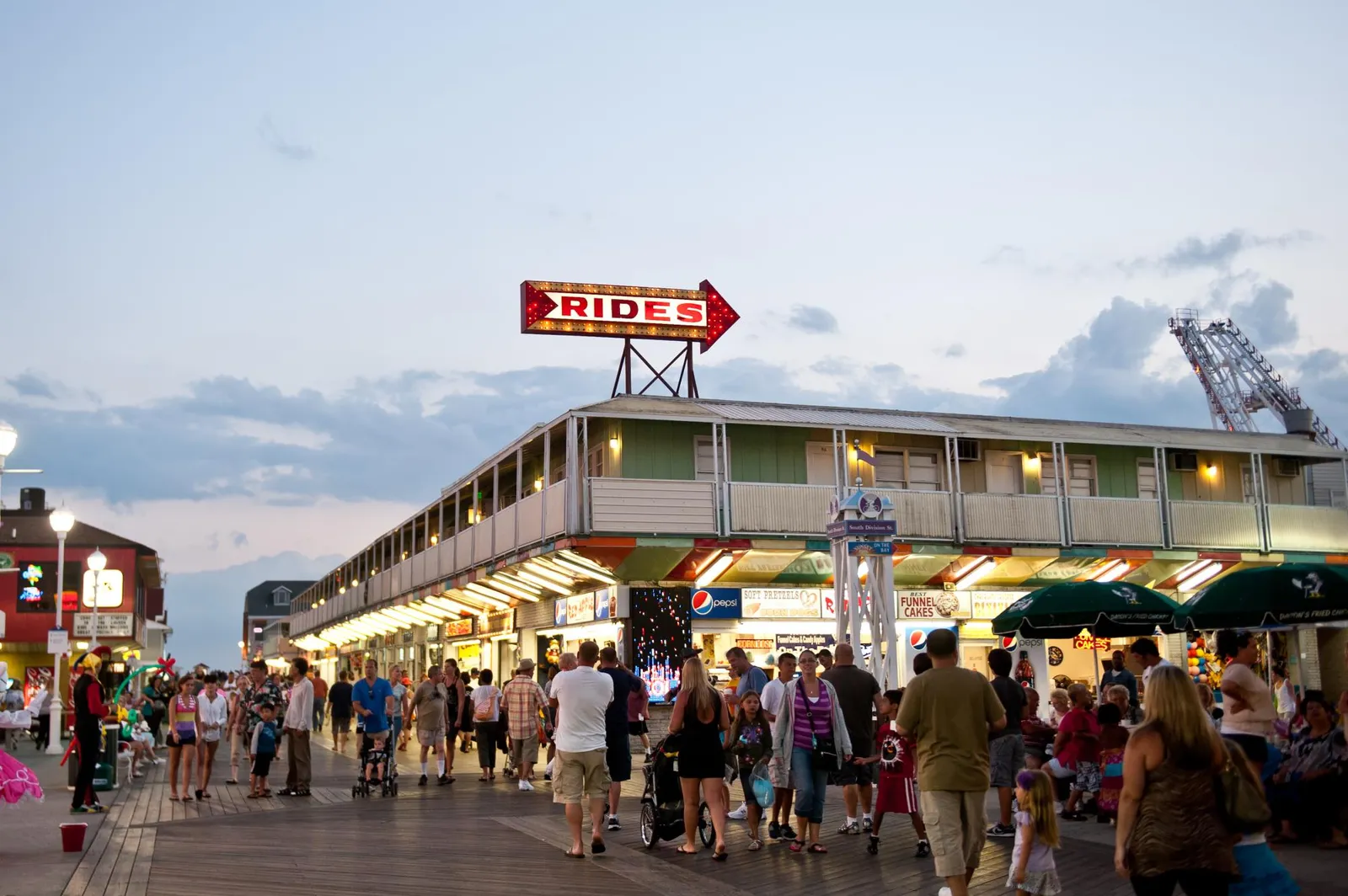 A crowded boardwalk scene at dusk with shops and a 'Rides' sign, suggesting a lively atmosphere.
