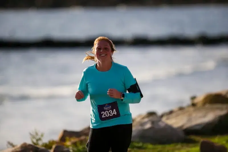 A female runner with red hair smiles as she runs along a waterfront path, wearing a teal long-sleeved shirt and black shorts.