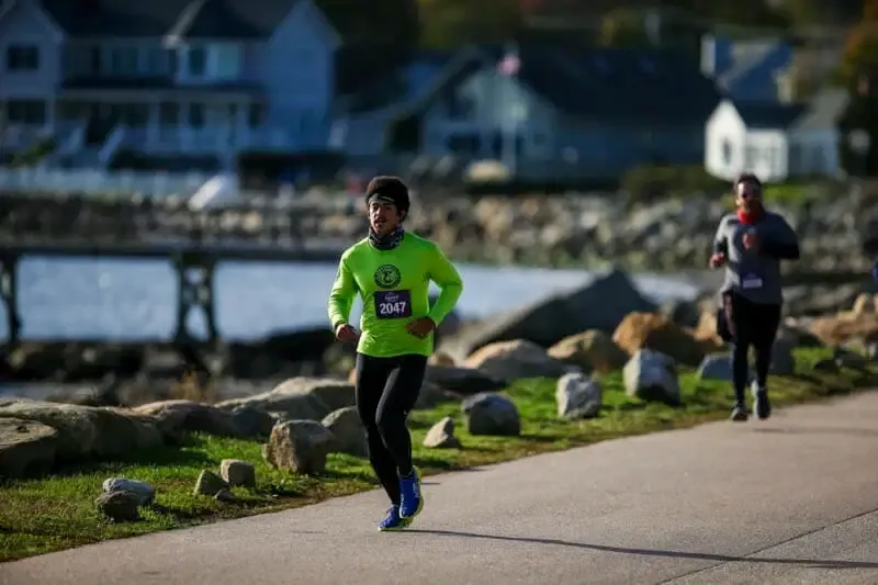 A runner in a bright green long-sleeved shirt with bib number 2047 runs along a paved path next to a body of water, with houses in the background.