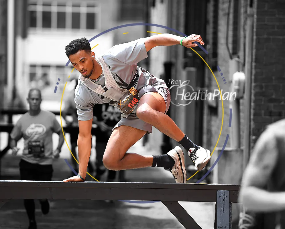 A male runner leaps over an obstacle during a race, with other runners blurred in the background.