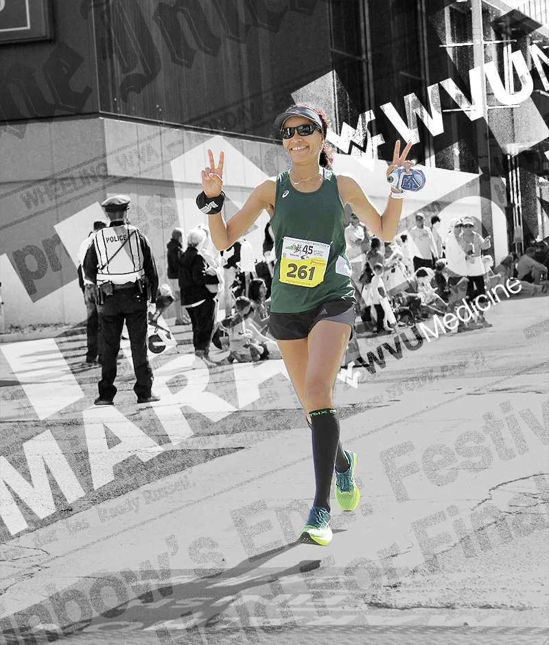 A female runner smiles and holds up two peace signs while running in a race, with spectators and a police officer in the background.