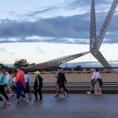 Runners crossing a bridge with a large, abstract metal sculpture overhead.