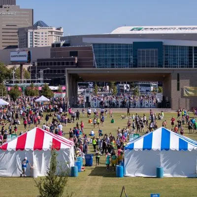 A finish line festival with many people gathered around tents and a stage, with city buildings in the background.