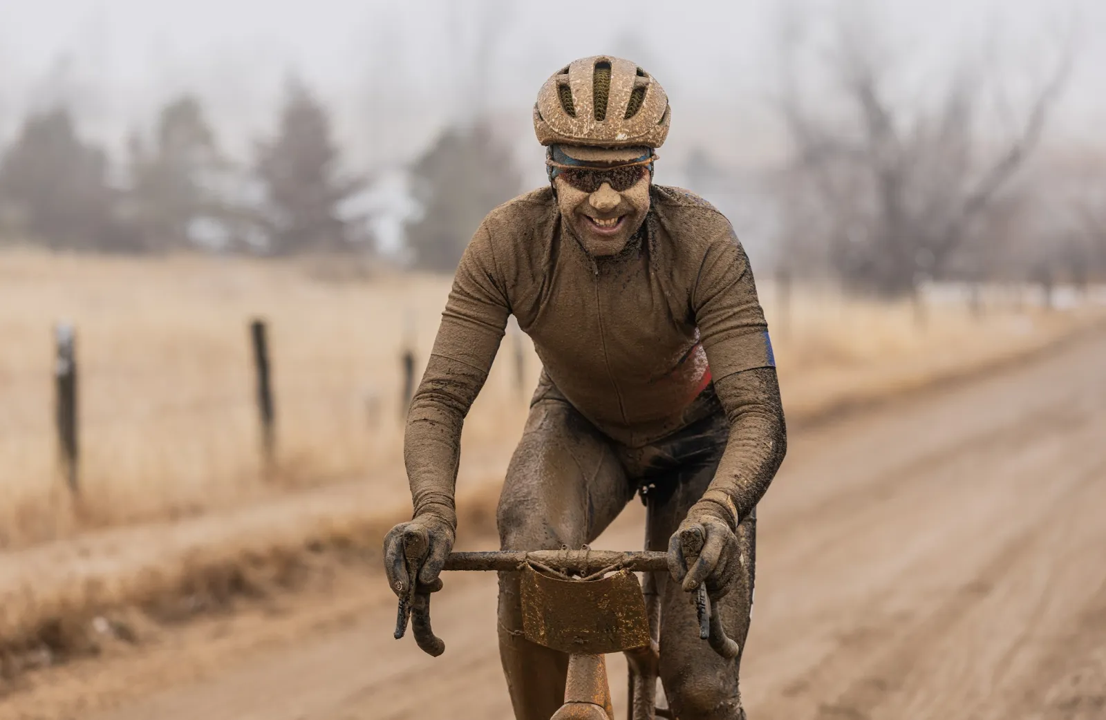 A cyclist covered head-to-toe in mud, smiling broadly as they ride on a dirt road during the Old Man Winter Rally.