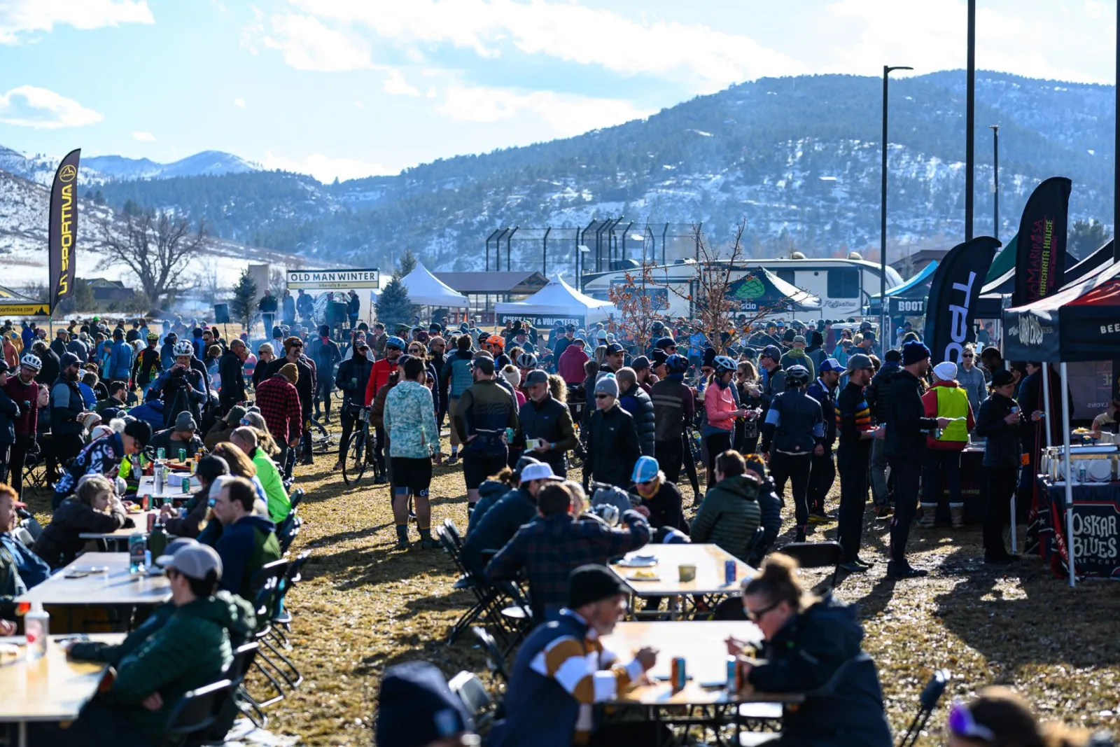 A large crowd of people gathered at an outdoor event, with tents and banners visible, under a bright sky with mountains in the background.