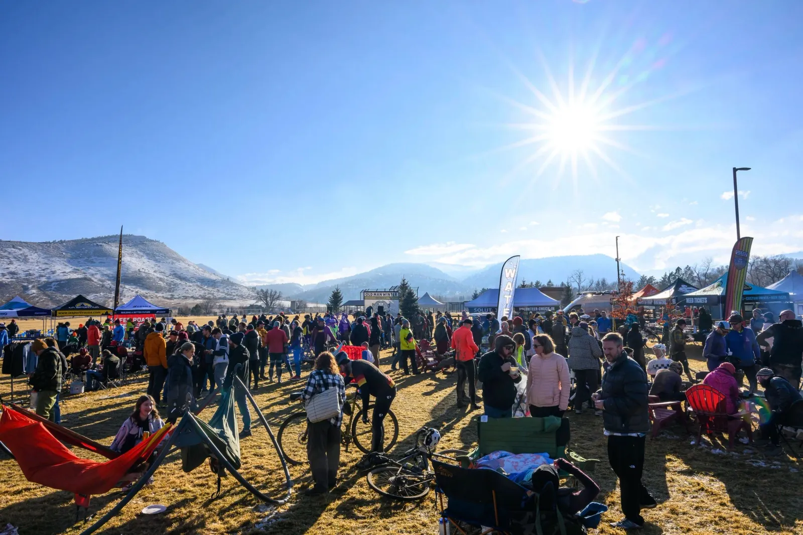 A crowd of people gathered at an outdoor event with tents and mountains in the background under a bright sun.