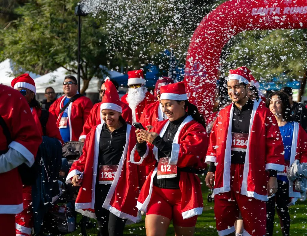 Runners dressed as Santa Claus smiling and running, with artificial snow falling around them.