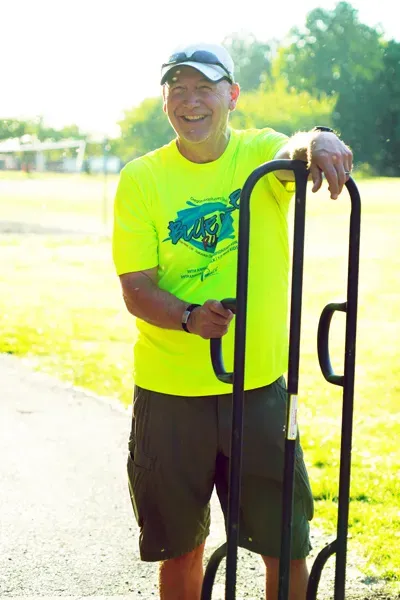 A smiling man wearing a bright yellow t-shirt and a white baseball cap, leaning on a metal cart.