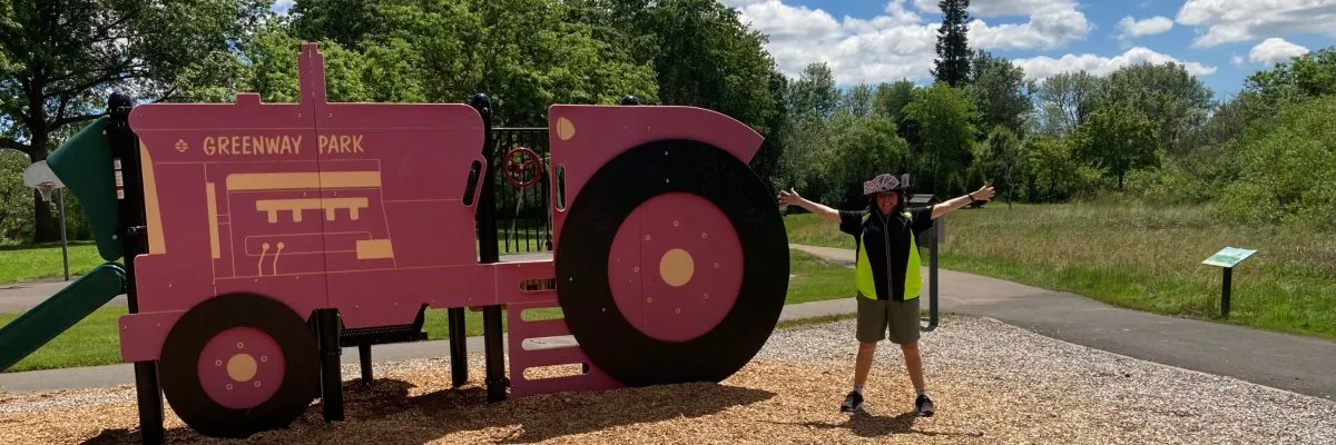 A person wearing a reflective vest and a hat decorated with playing cards stands with arms outstretched in front of a large pink tractor structure in a park.