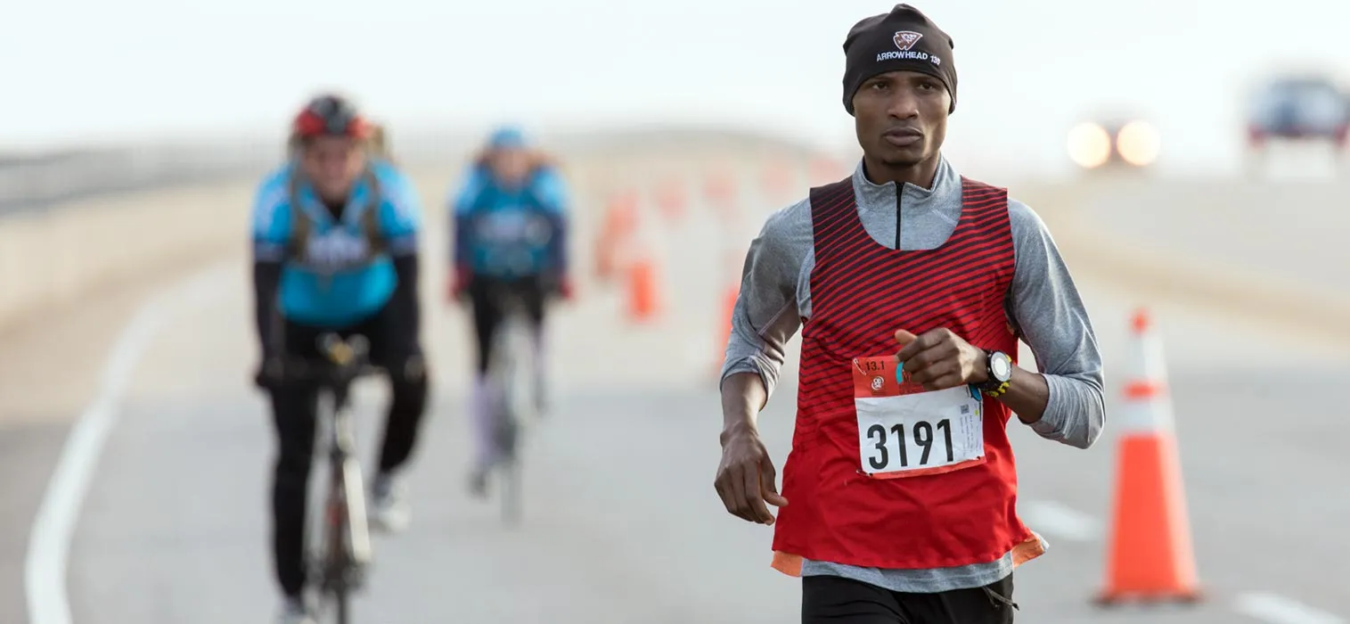 A male runner wearing a red and black striped singlet and a black beanie runs on a road, with other cyclists blurred in the background.