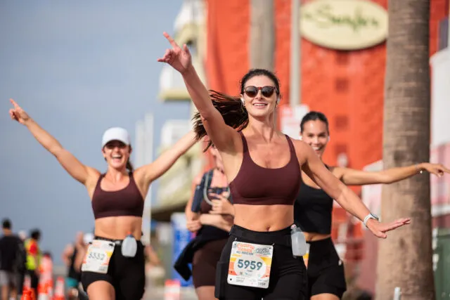 Three women running a half marathon, with the lead runner in the foreground smiling and giving a peace sign, appearing to be near the finish line.