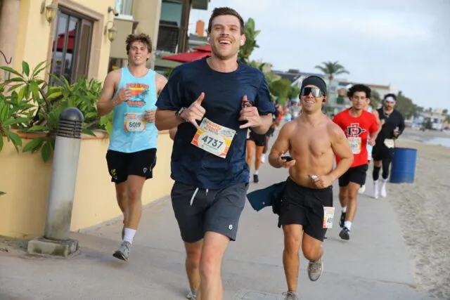 Runners smiling and giving thumbs up as they race along a beach path.