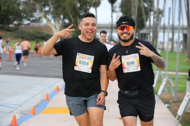 Two male runners smiling and giving peace signs at a race, with other runners in the background.
