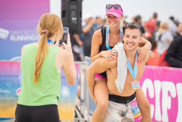 A man gives a woman a piggyback ride, both wearing race medals and smiling, with a crowd in the background.