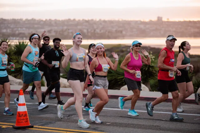 A diverse group of runners smiling and waving as they run along a road with a scenic background of water and hills.