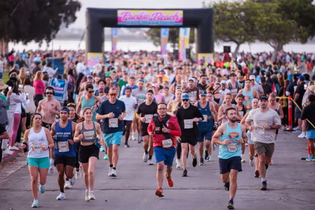 A large group of runners starting a half marathon under a colorful archway that says 'Pacific Beach Half Marathon'.