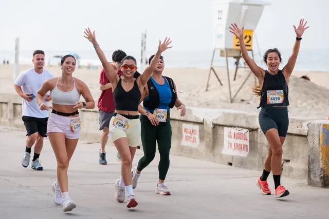 A group of diverse runners with their arms raised in celebration during a beachside half marathon.
