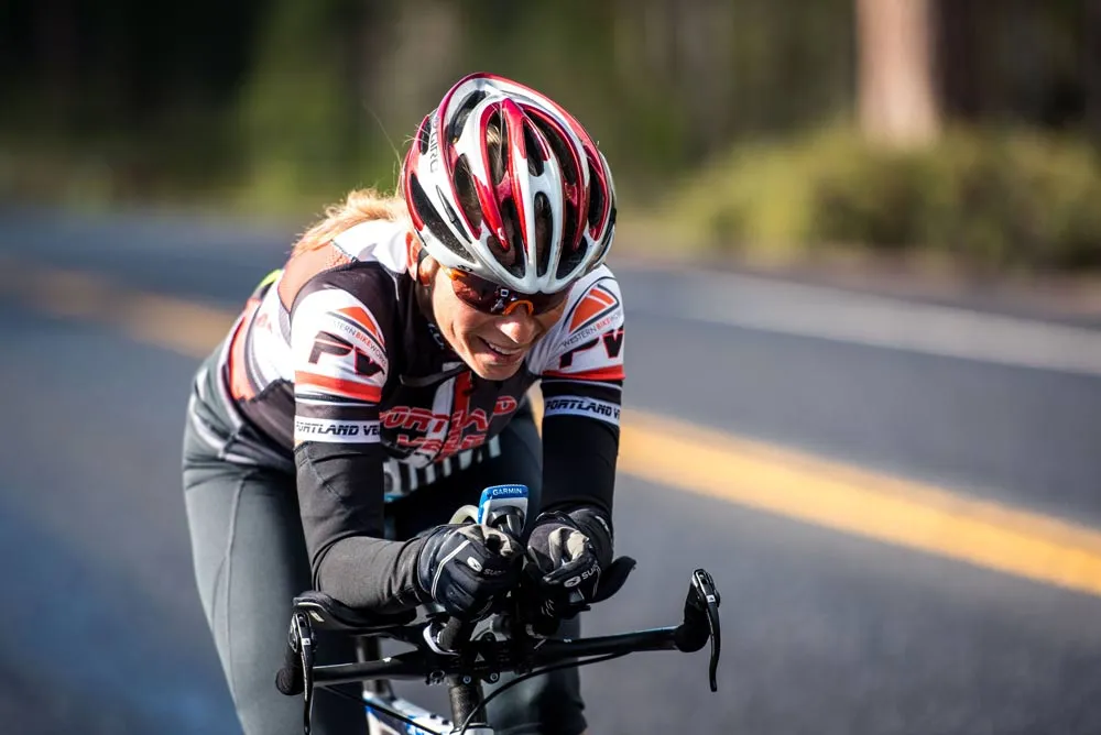A female cyclist in a red and white helmet and racing jersey rides on a road with a yellow line.