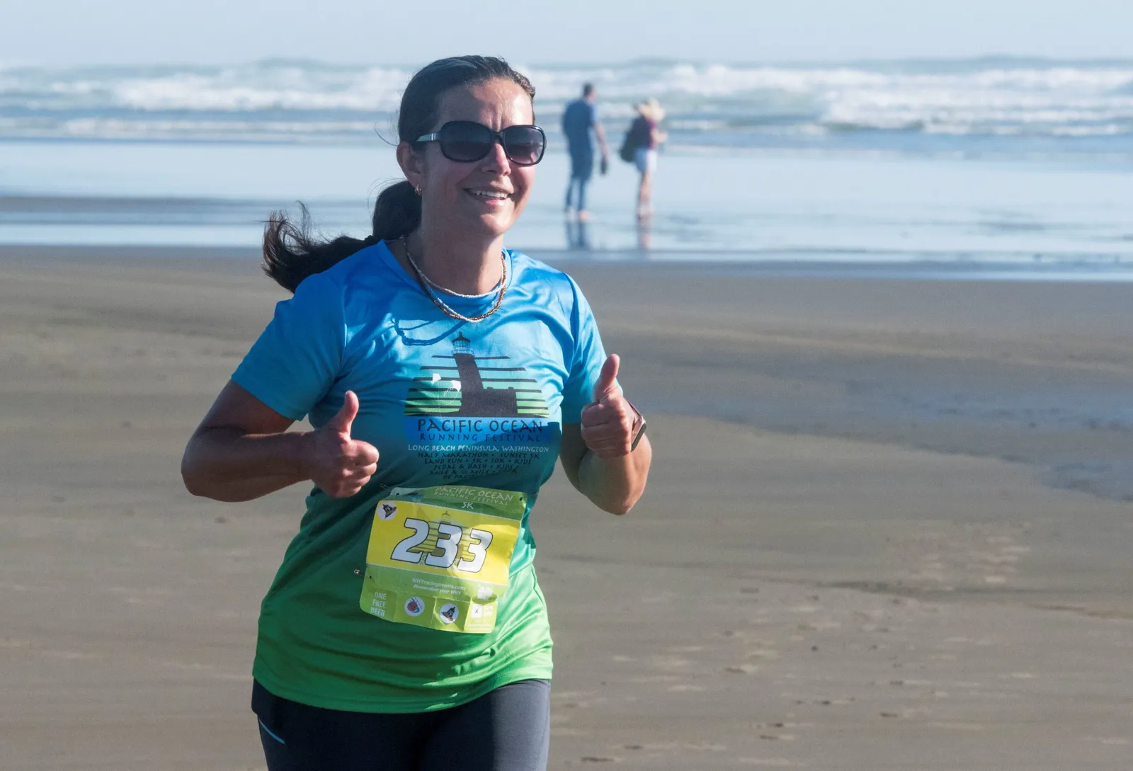 A female runner smiles and gives two thumbs up while running on a beach during the Pacific Ocean Running Festival.