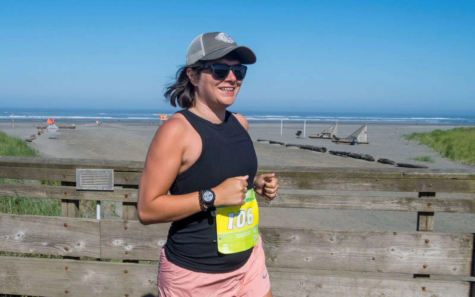 A woman wearing a black tank top, pink shorts, sunglasses, and a baseball cap runs along a wooden boardwalk with the ocean and beach in the background.
