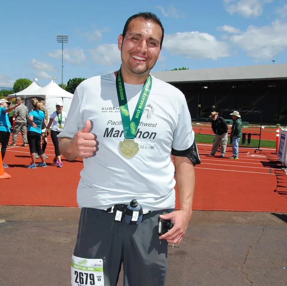 A smiling runner gives a thumbs up while wearing a medal and a race t-shirt.