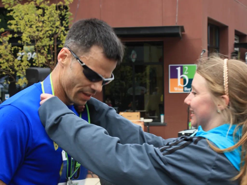 A runner with sunglasses and a blue shirt is receiving a medal from a smiling woman.
