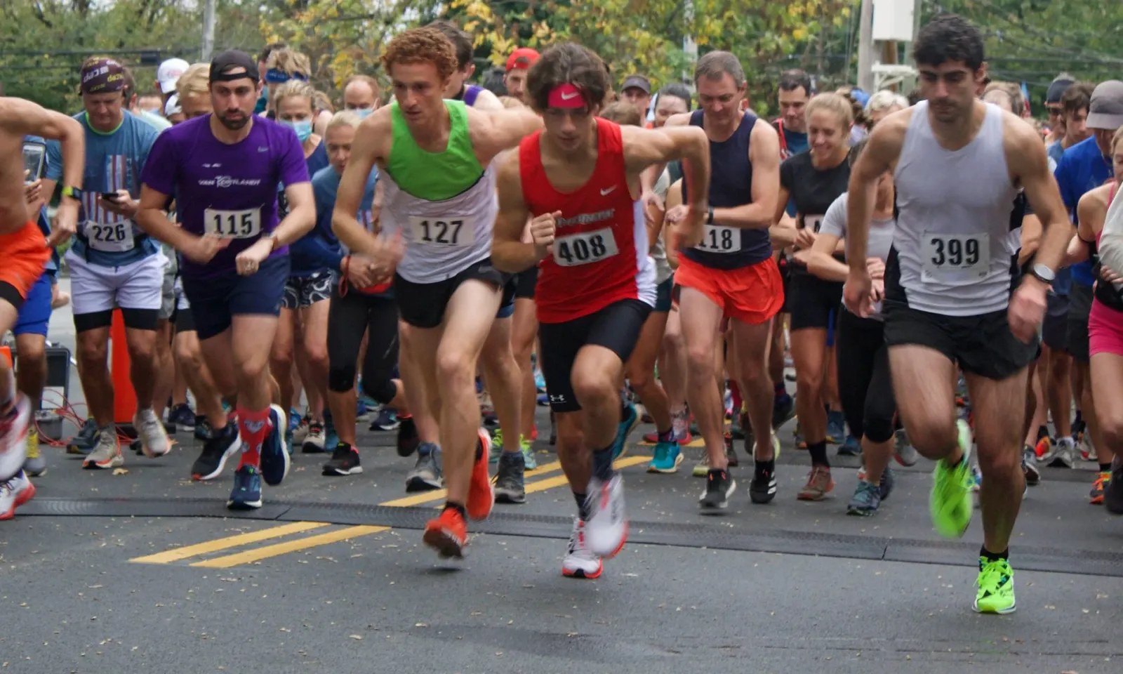 A group of male runners in the foreground, starting a race on a paved road, with other runners and spectators in the background.