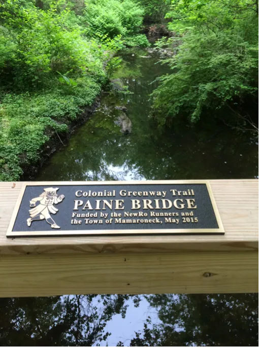 Plaque for Paine Bridge on the Colonial Greenway Trail, funded by NewRo Runners, with a scenic view of the wooded trail and creek in the background.
