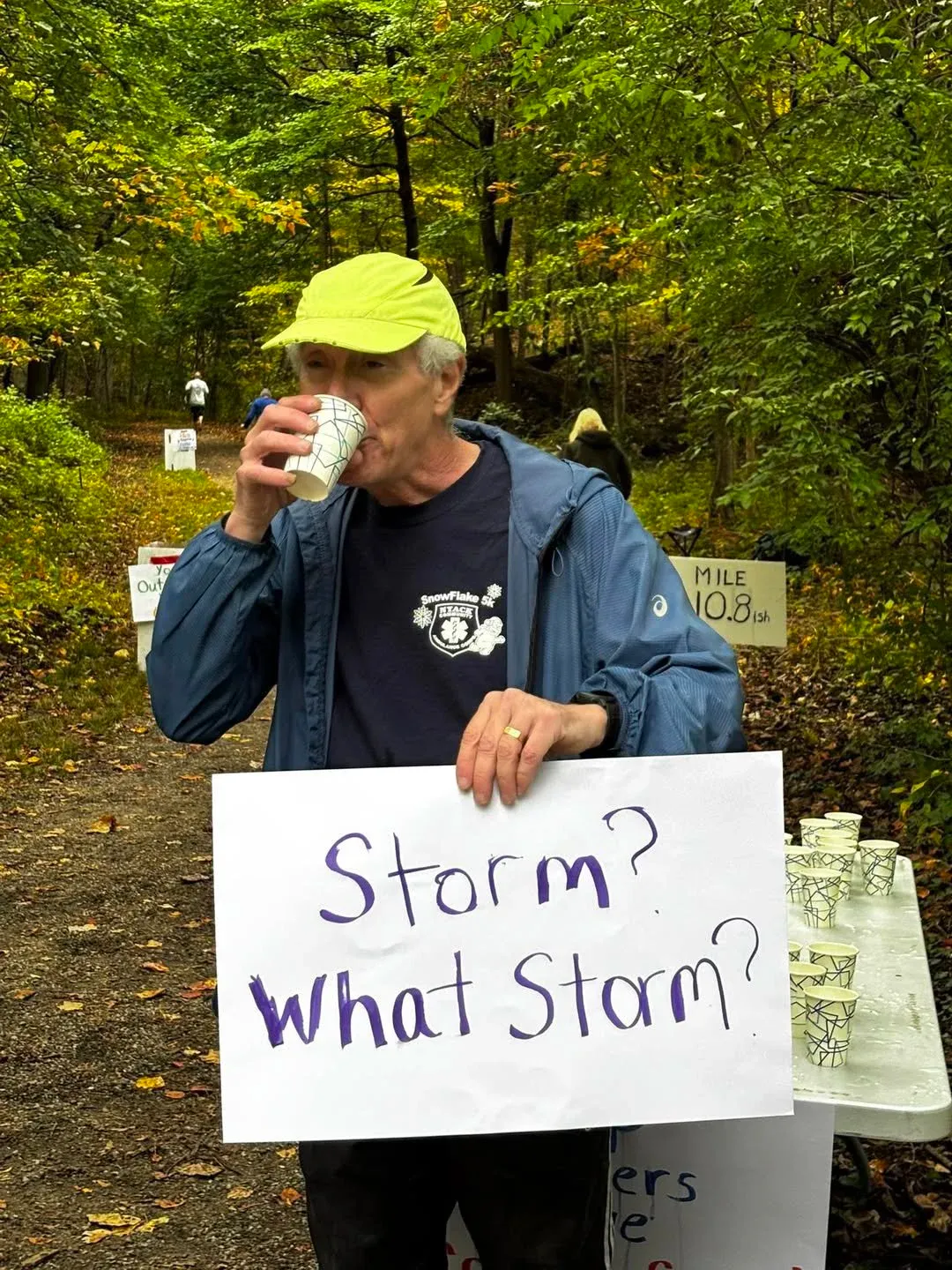 A runner drinks from a cup at an aid station, holding a sign that reads 'Storm? What Storm?' with other runners and mile markers visible in the background.