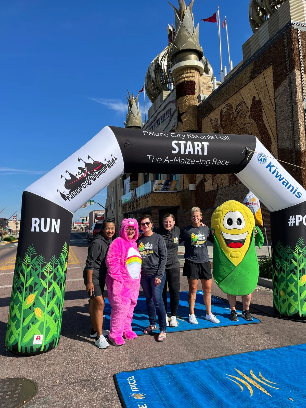 A group of people, including one in a pink costume and another in a corn mascot costume, pose under a "START" archway at the Palace City Kiwanis Half Marathon.