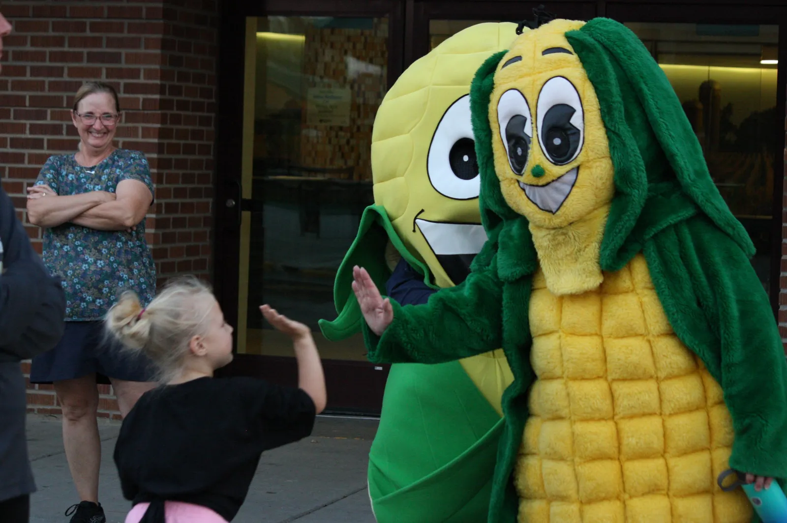 A child waves at a person in a corn mascot costume, with another mascot behind them and a woman smiling in the background.