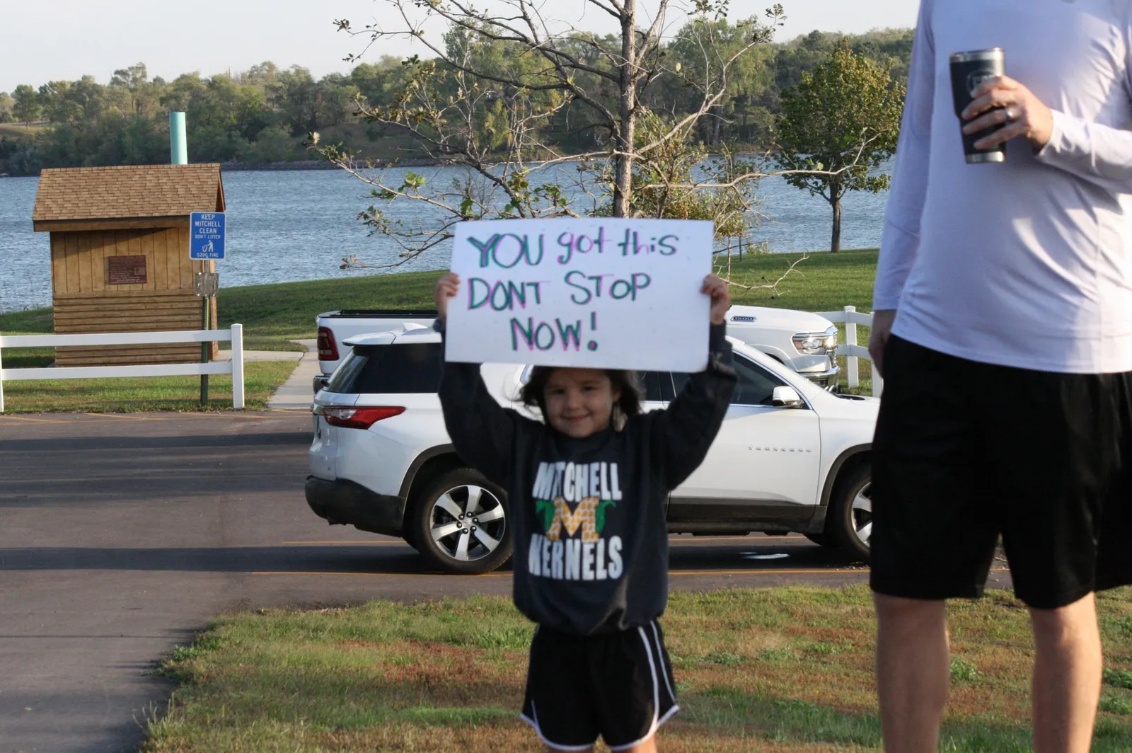 A child holds up a sign that says "YOU got this DONT STOP NOW!" with a runner in the foreground and a lake in the background.