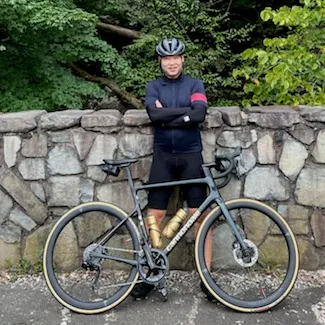 A man in cycling gear stands with his road bike next to a stone wall, with trees in the background.