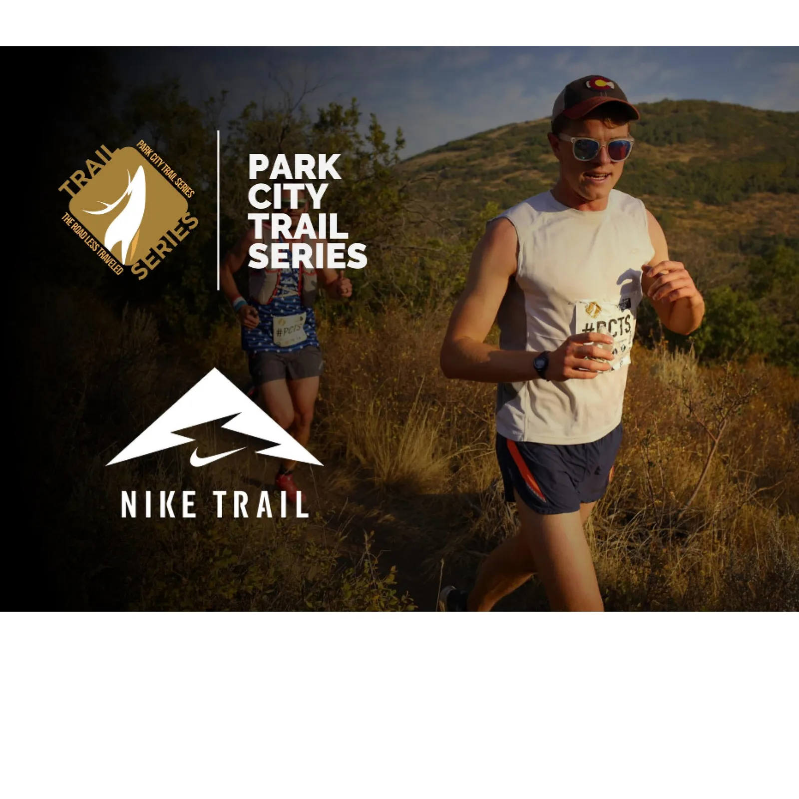 A male runner smiles as he runs on a trail, with another runner in the background and mountains in the distance.