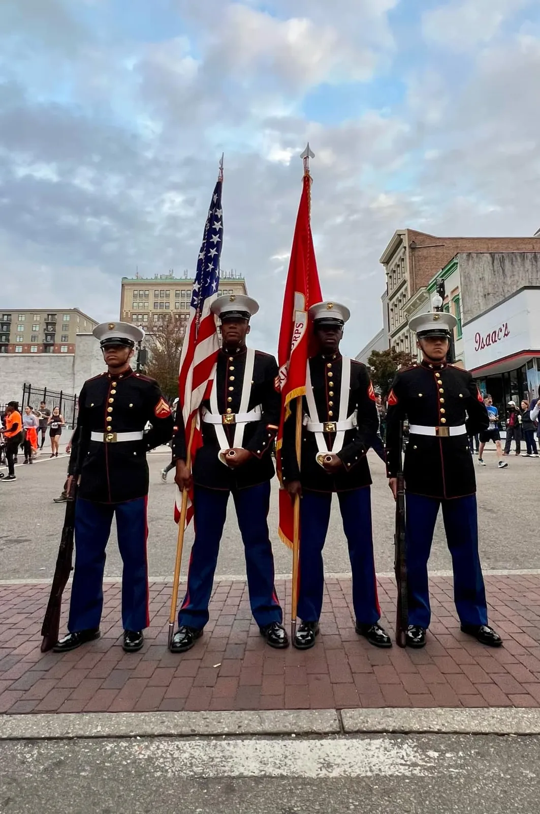 Four uniformed individuals stand with American and Marine Corps flags, with runners visible in the background.