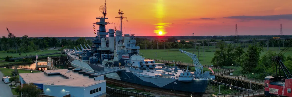 Battleship docked at sunset with a vibrant orange and yellow sky.