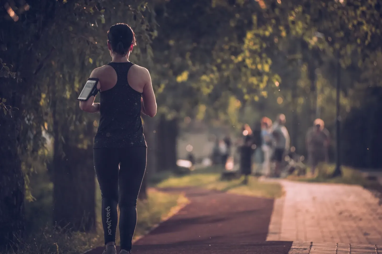 A woman runs on a paved path in a park, with trees and dappled sunlight.