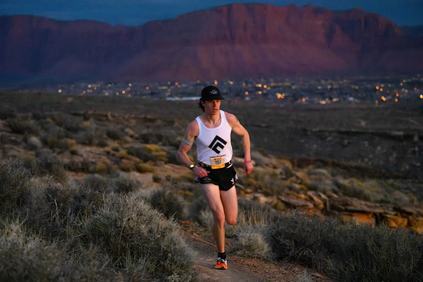 A male runner with bib number 103 runs on a dusty trail at dusk, with a city skyline and mountains in the background.