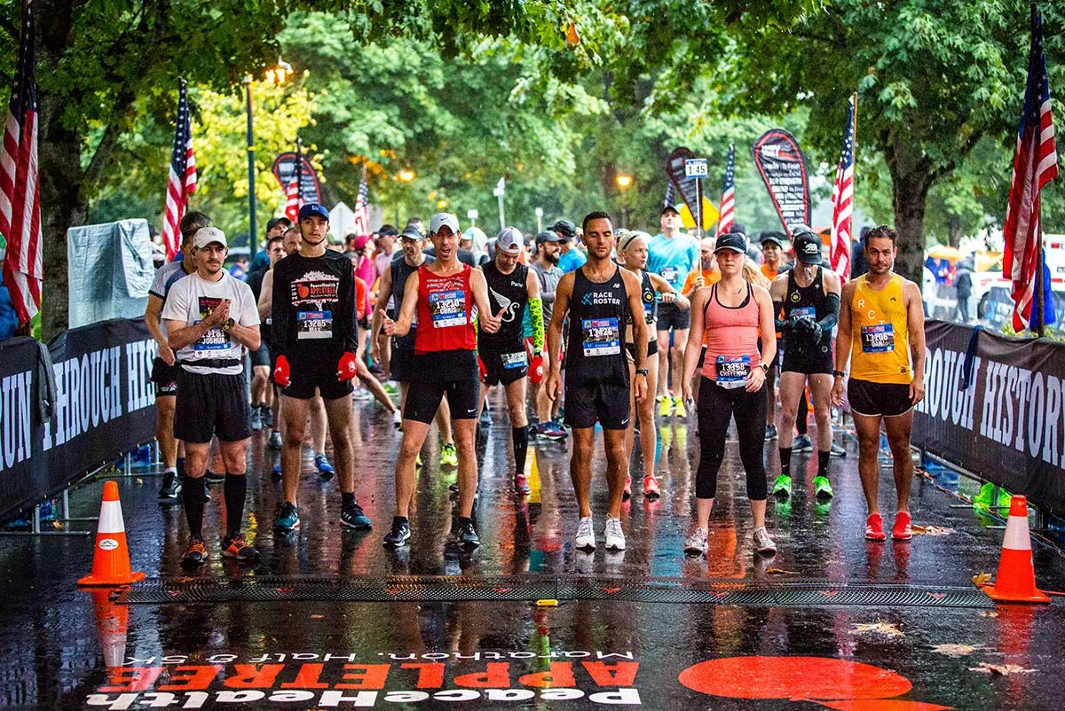 Runners lined up at the start of a race, with American flags and banners in the background, on a wet road.