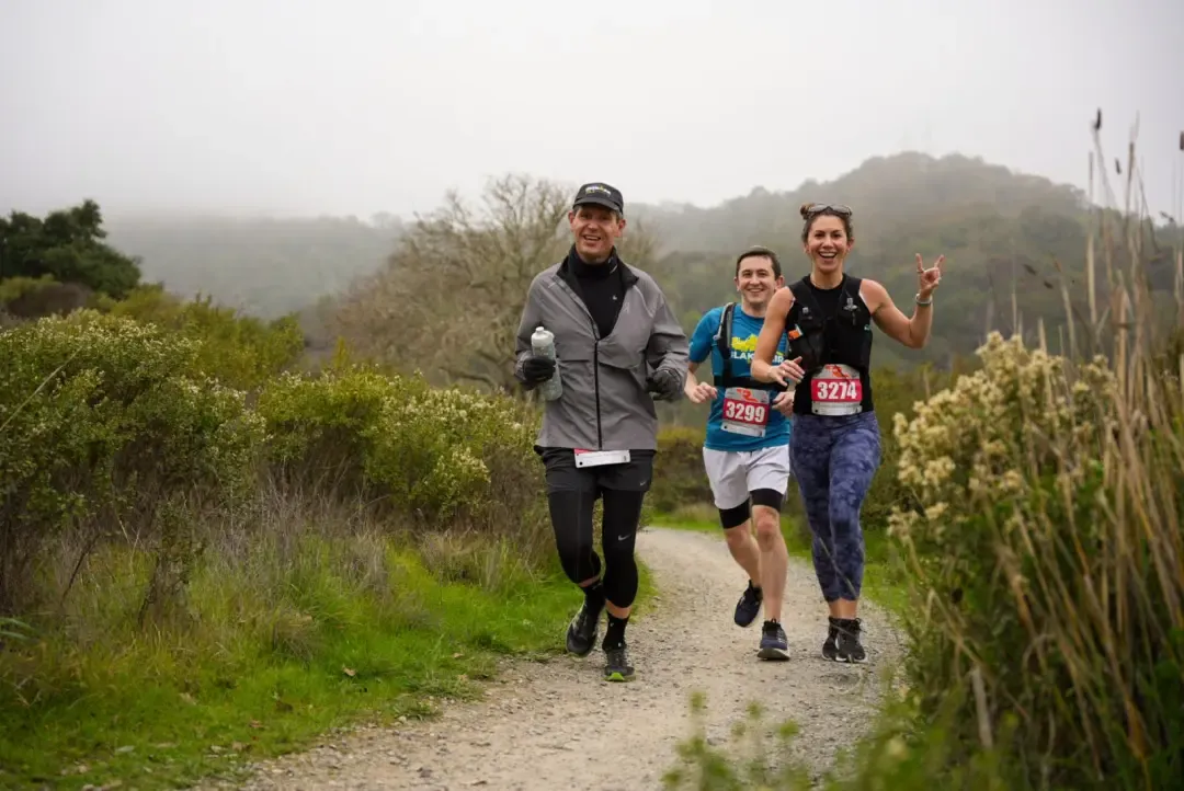 Three runners smiling and running on a dirt path with green foliage and hills in the background.