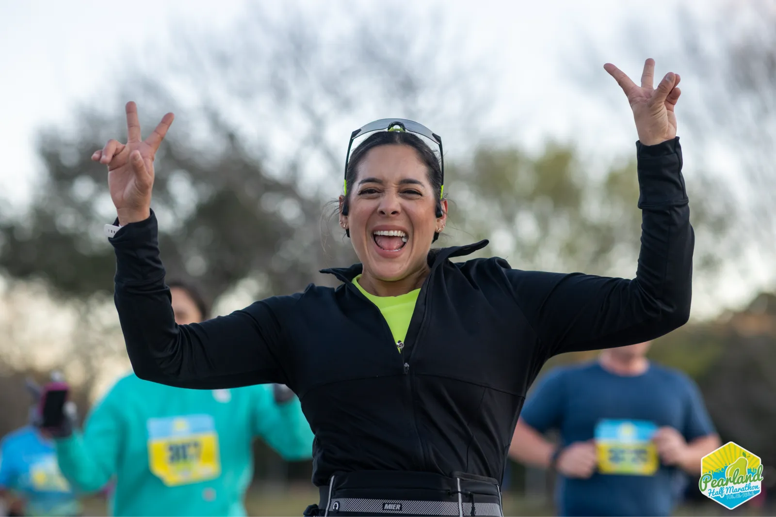 A woman with her arms raised in a V-sign gesture, smiling broadly and shouting with excitement during a half marathon.