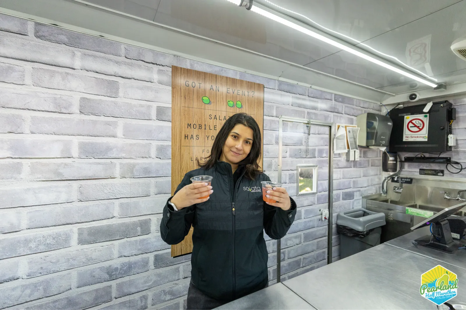A woman in a black jacket holding two cups of drinks, smiling at the camera, with a sign behind her that says 'GOT AN EVENT?'
