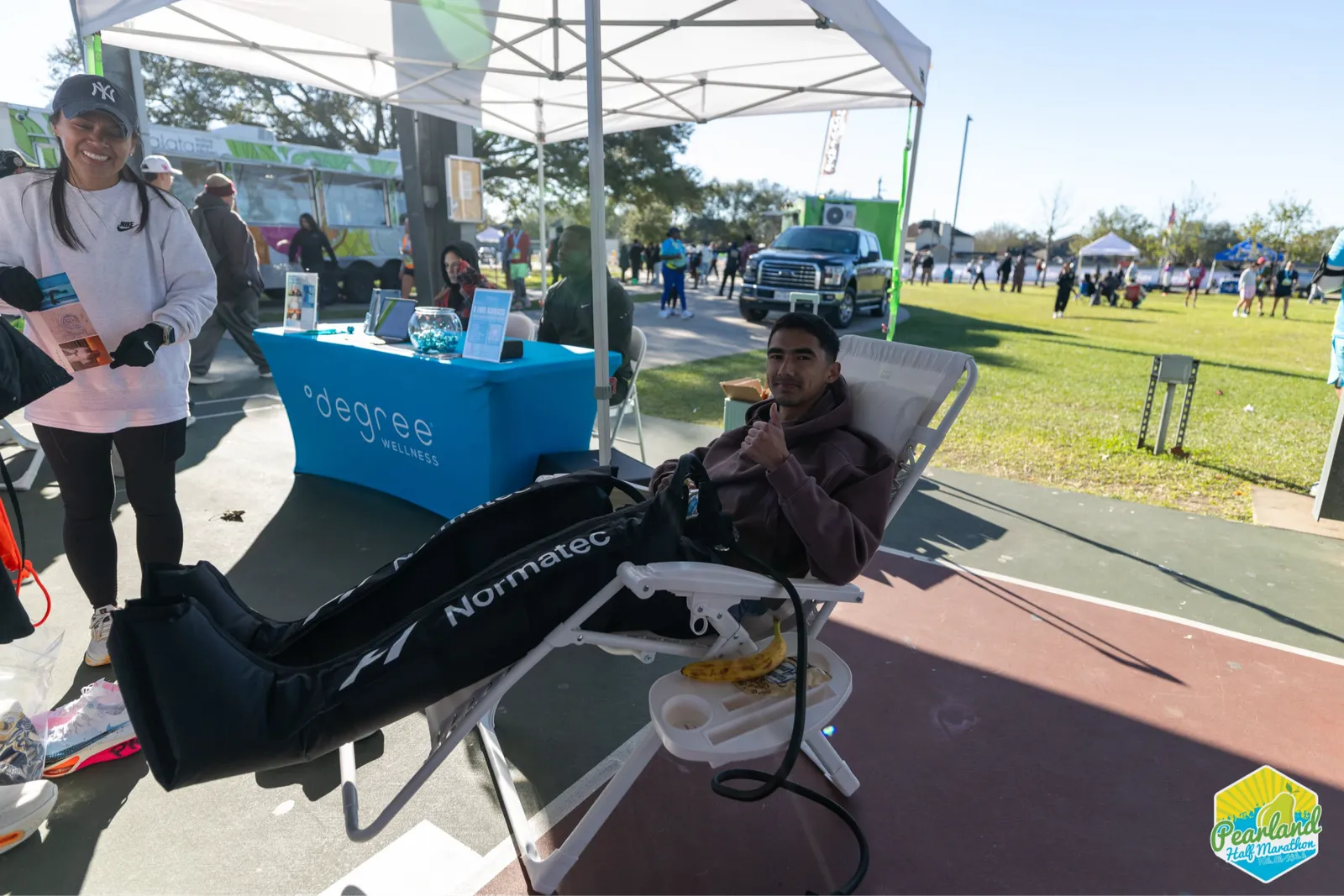 A man gives a thumbs up while receiving leg compression therapy at a post-race recovery tent, with other participants and tents visible in the background.
