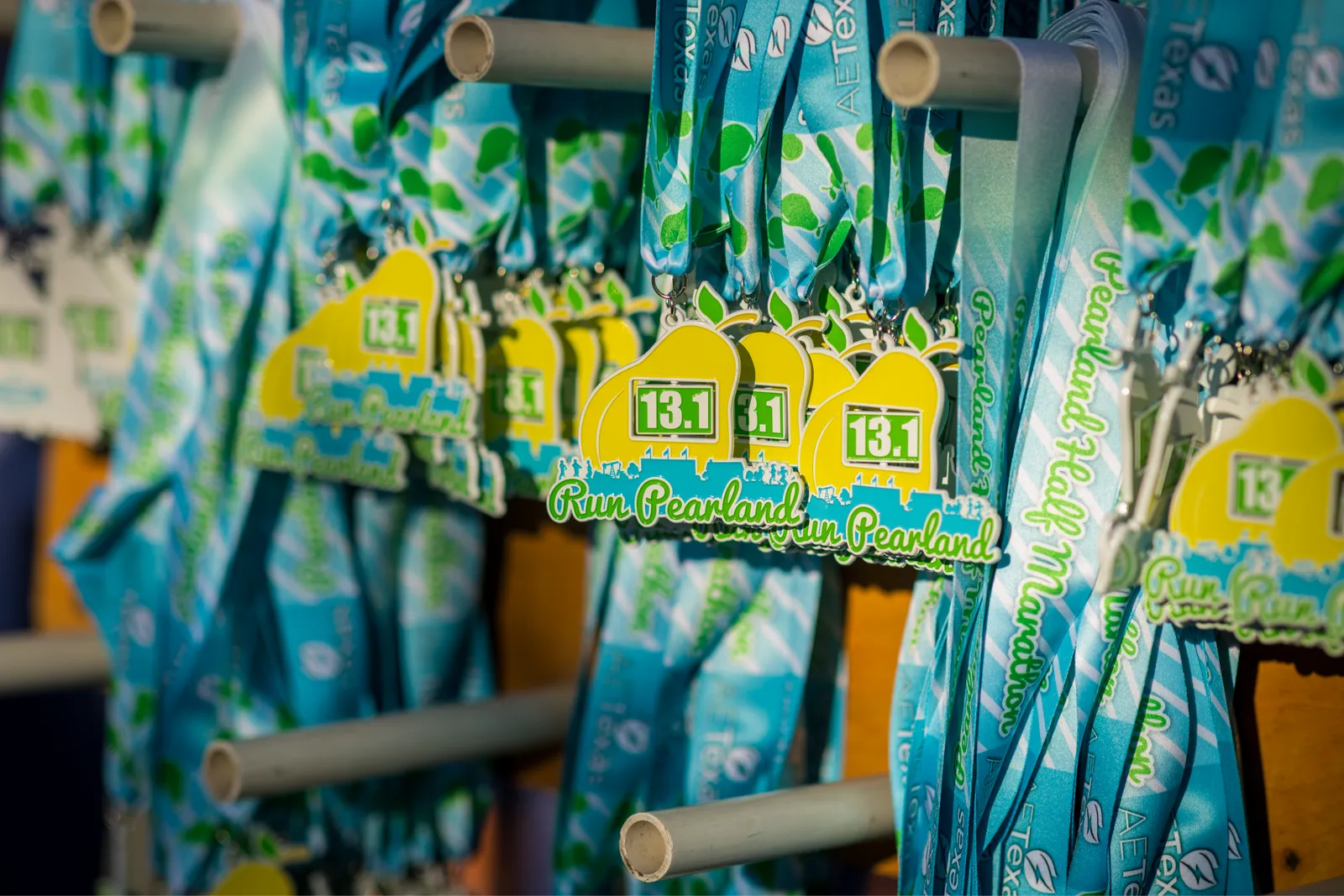 Close-up of yellow pear-shaped medals with '13.1 Run Pearland' text and blue ribbons with green leaf patterns hanging on a rack.