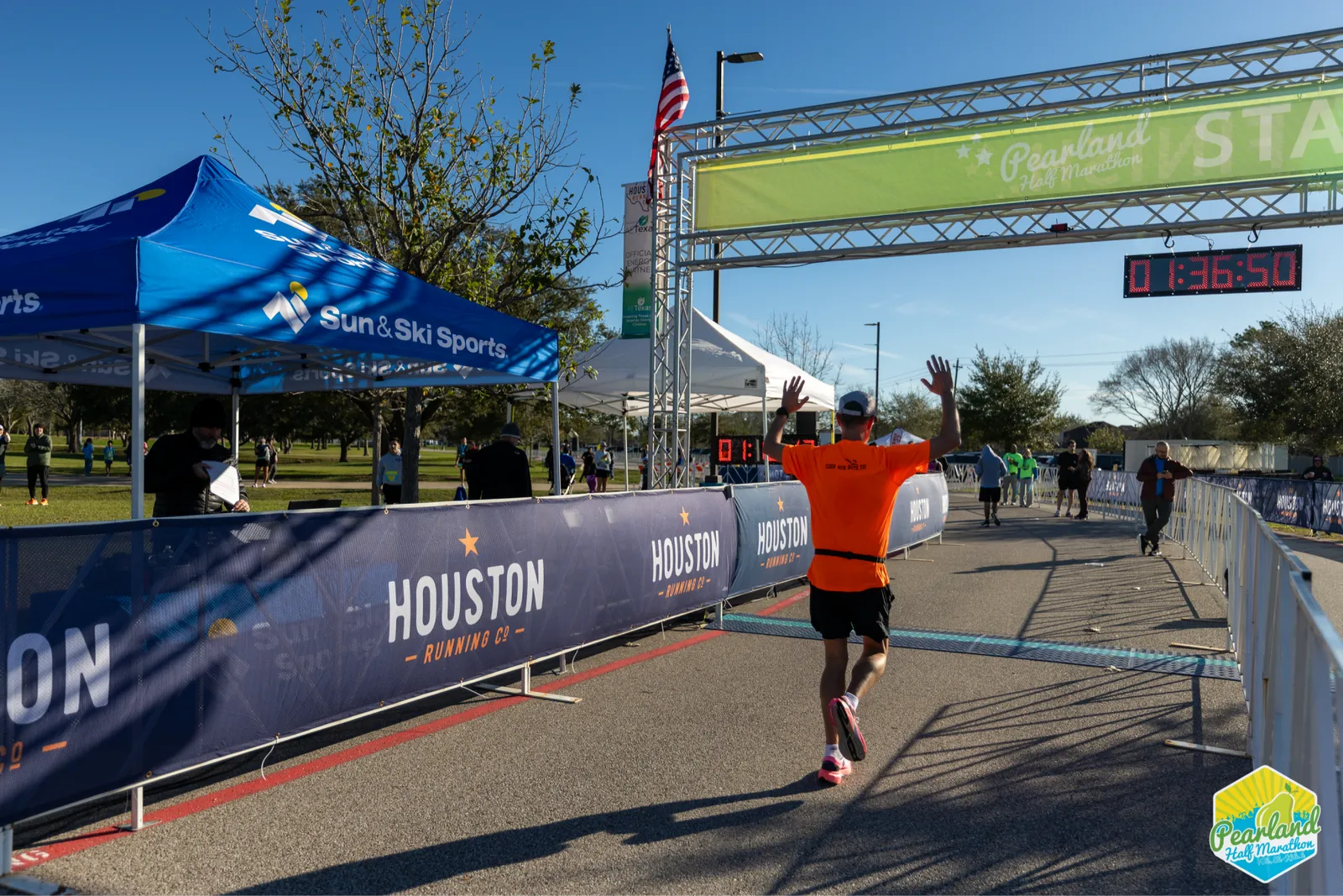 A runner in an orange shirt crosses the start/finish line with arms raised in celebration at the Pearland Half Marathon.