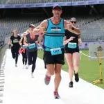 A male runner in a blue tank top and black shorts leads a group of runners on a paved path, with stadium seating in the background.