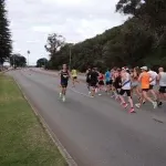 Runners participating in the Perth Half Marathon, running along a paved road with trees and a hill in the background.
